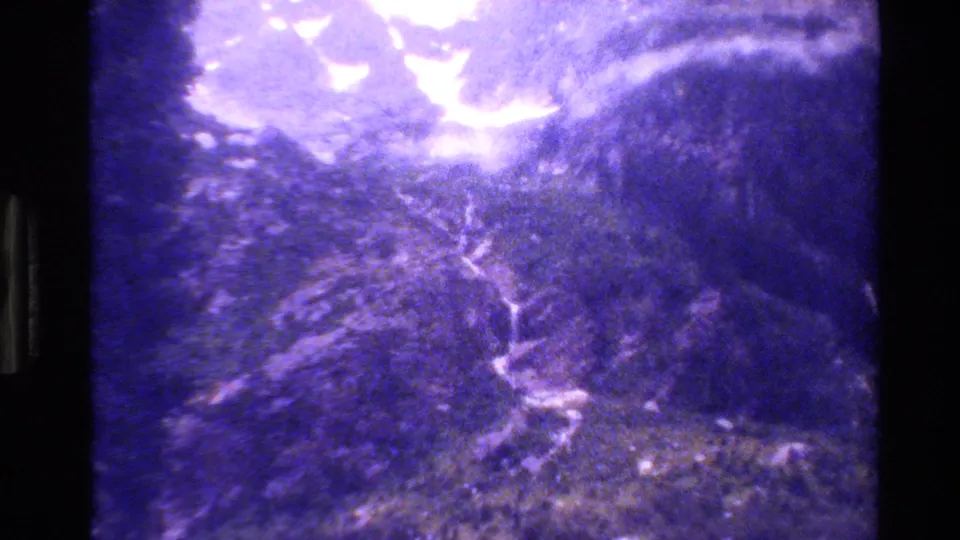 1980 Grand Teton 8mm frame showing rugged forested mountain terrain, bright overexposed sky, and a pale winding waterfall or path through the slope.