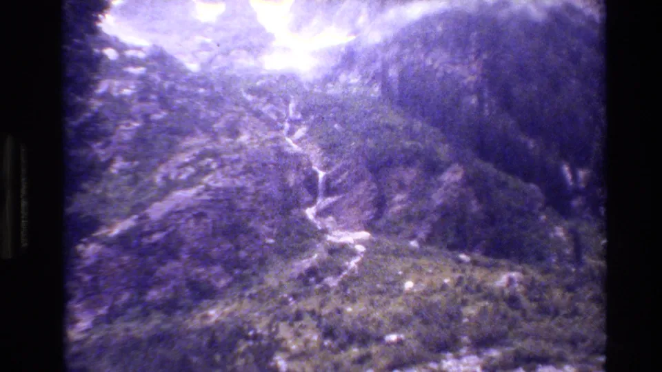 1980 Grand Teton home-movie frame showing steep wooded slopes, a bright sky area, and a light waterfall or drainage line descending through the landscape.