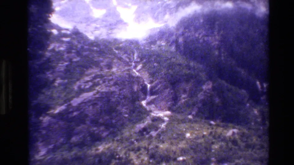 1980 Grand Teton 8mm frame showing a grainy elevated view of forested mountain slopes, a pale winding waterfall or trail, and dark scan borders.