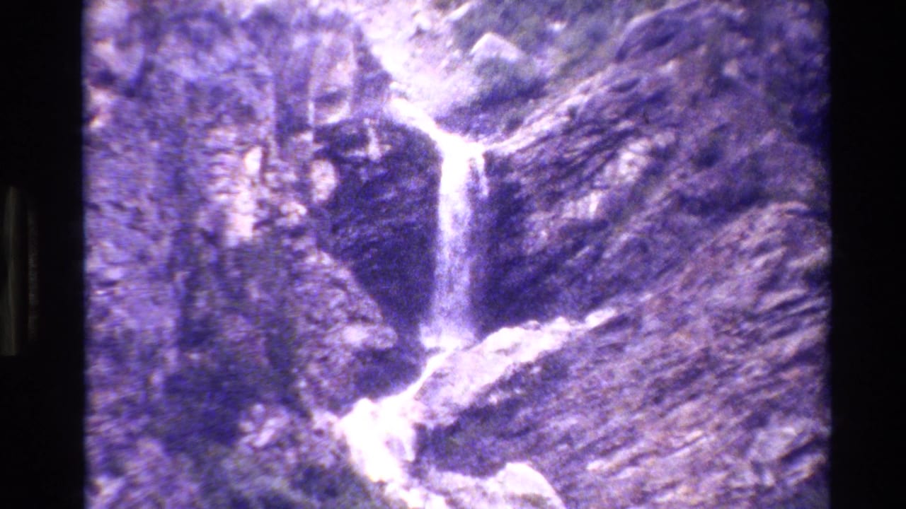 Grand Teton home-movie frame with a narrow waterfall descending over layered rock in a purple-faded 8mm scan.