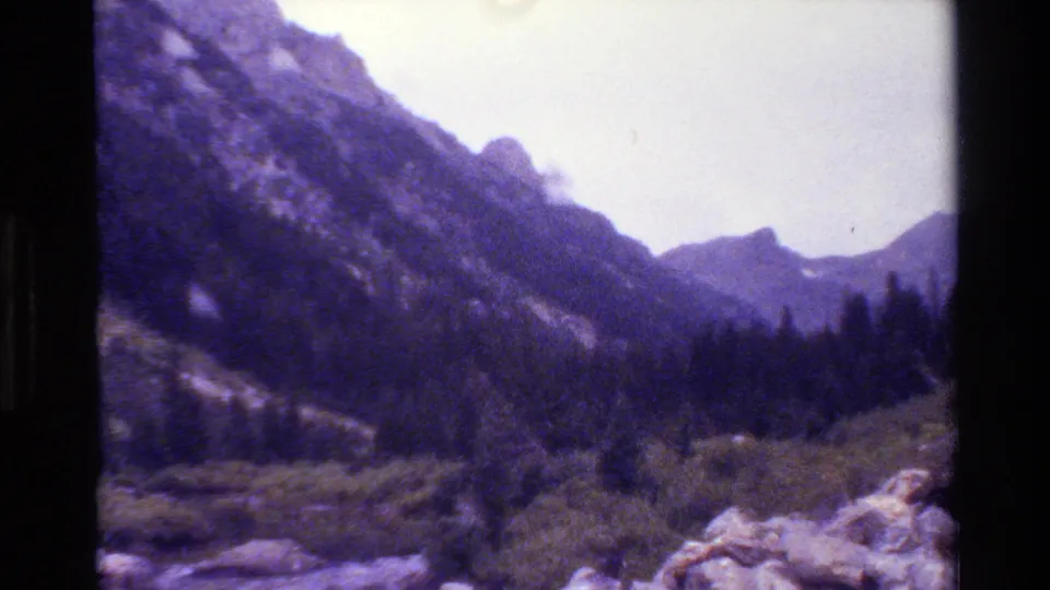 1980 Grand Teton 8mm frame with rocky mountain slope, distant peaks, evergreen valley, and purple-blue film color shift.