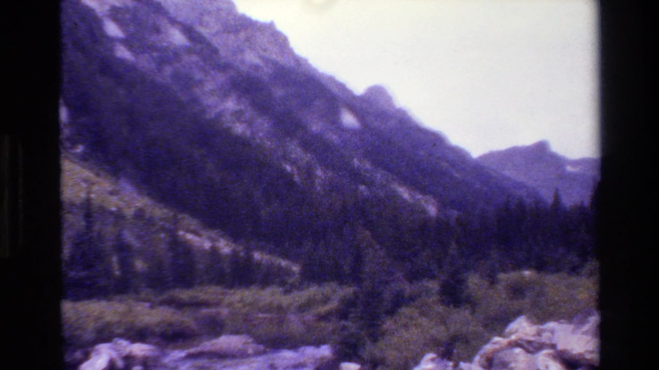 1980 8mm home-movie view of Grand Teton mountain slopes with forested foothills, rocky foreground, and a pale sky.
