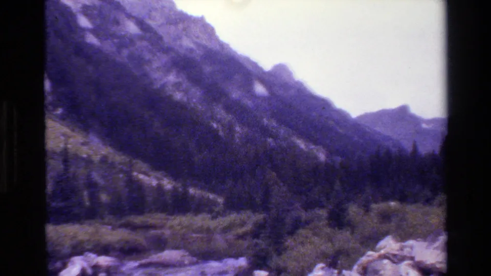 1980 8mm home-movie view of Grand Teton mountain slopes with forested foothills, rocky foreground, and a pale sky.