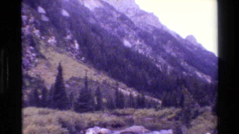 1980 Grand Teton home-movie frame with forested foothills, rocky foreground, and a purple-shifted 8mm film look.
