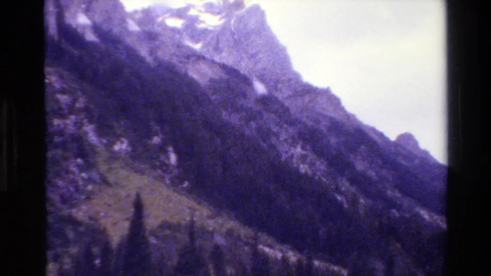 Grand Teton mountain view from 1980 8mm footage, with a snow-dusted peak, steep slope, forest, and bright sky.