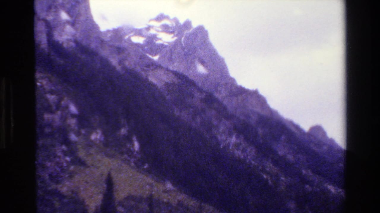 1980 Grand Teton home-movie frame showing jagged snow-capped peaks, rugged slopes, and cloudy sky with a purple 8mm color cast.