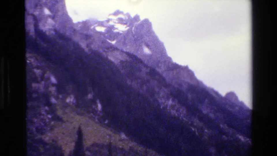 1980 Grand Teton home-movie frame showing jagged snow-capped peaks, rugged slopes, and cloudy sky with a purple 8mm color cast.