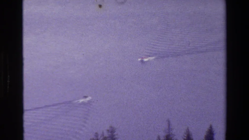 1980 Grand Teton lake scene with two small boats moving across calm water, visible wakes, and dark pine trees along the lower edge.
