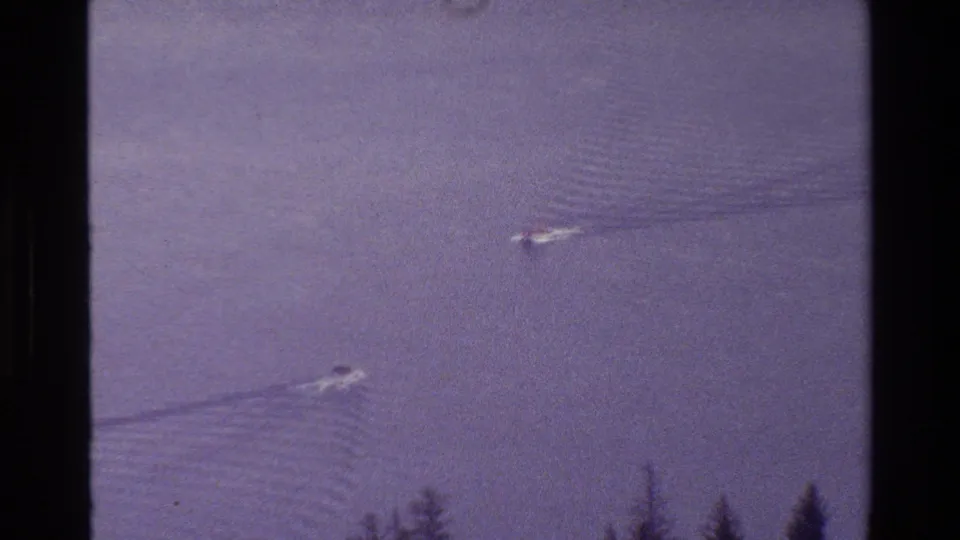 Grand Teton lake frame from 1980 showing two distant boats, rippled wakes, broad water surface, and shoreline trees in purple-tinted 8mm footage.