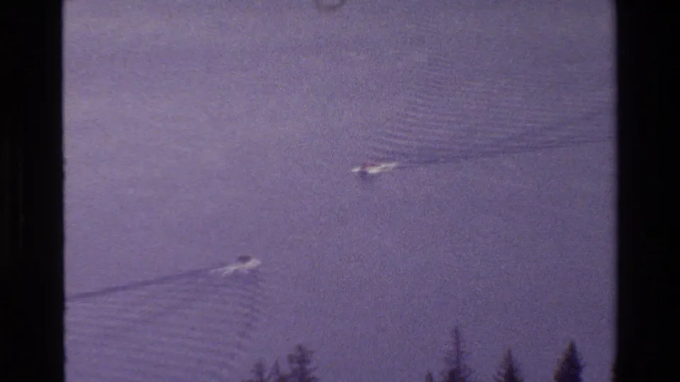 Grand Teton, Wyoming 1980 home-movie frame showing two small boats crossing calm lake water near an evergreen shoreline.