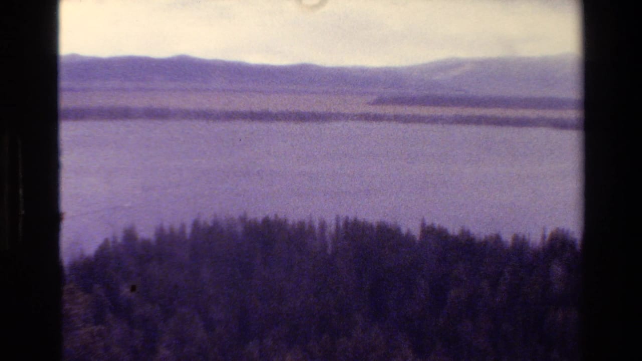 1980 8mm view in Grand Teton, Wyoming, showing a broad lake, evergreen foreground, and distant mountain ridge under an overcast sky.