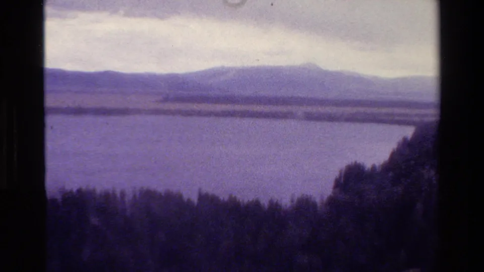 Visitor-shot 1980 Grand Teton frame looking over a lake toward muted mountain ridges, with dark forest in the foreground.