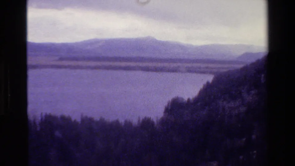 8mm Grand Teton landscape view with lake, distant mountain range, overcast sky, and a dark forested hillside entering from the right.