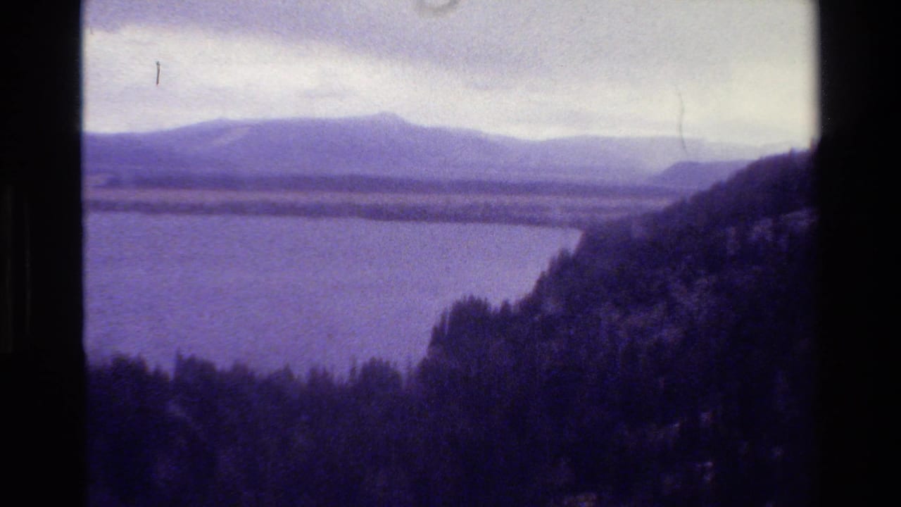 1980 8mm Grand Teton, Wyoming view of a lake bordered by dark forested hills beneath a cloudy sky.
