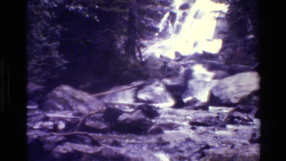 Vintage 8mm frame from Grand Teton shows a bright waterfall area above rocks, logs, and fast-moving water.