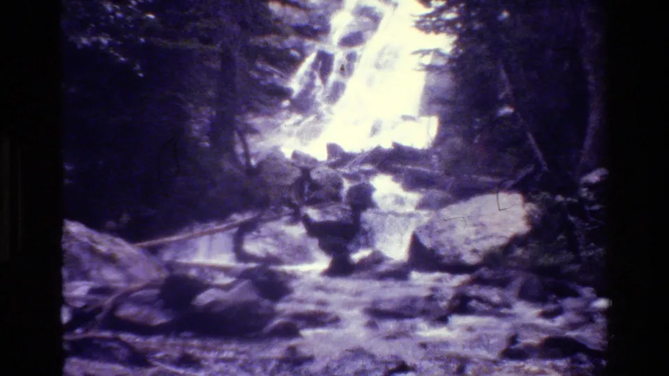 Grand Teton 8mm footage holds on a waterfall, rocks, logs, and shadowed trees along a rushing mountain river.