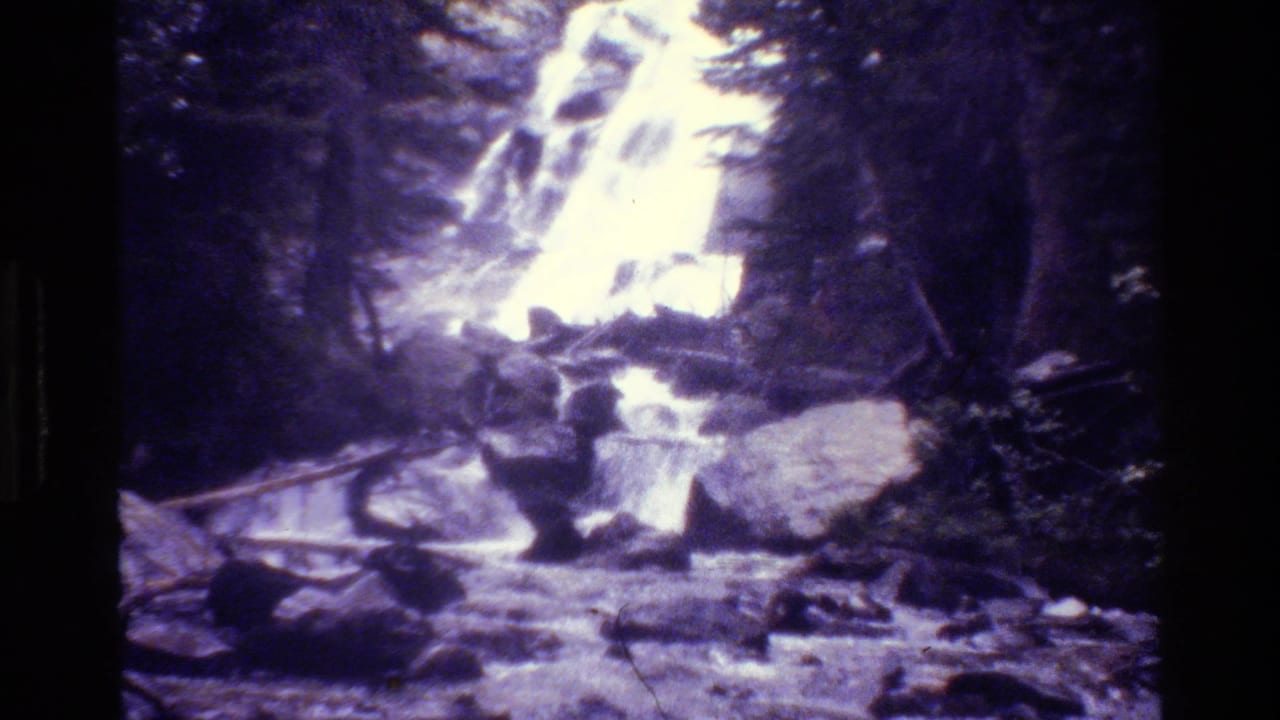 1980 Grand Teton 8mm frame of a bright forest waterfall surrounded by evergreen trees and rough rocks.