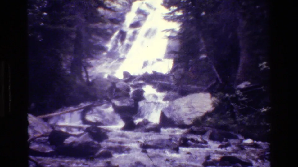 1980 Grand Teton 8mm frame of a bright forest waterfall surrounded by evergreen trees and rough rocks.