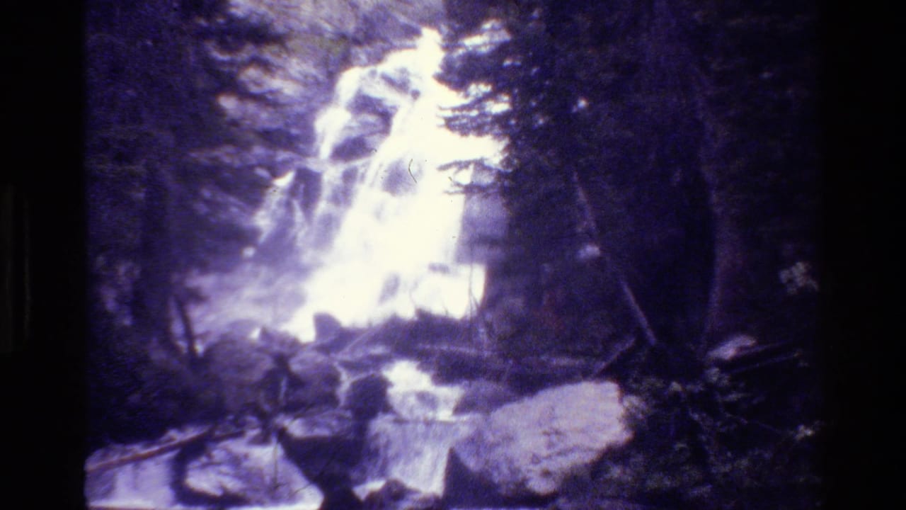 Vintage 8mm view of a Grand Teton waterfall seen through trees, with rocks and shaded forest around it.