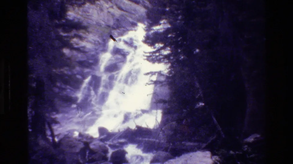 Purple-cast 1980 home-movie frame showing a bright waterfall, dark evergreens, and rocky terrain in Grand Teton.