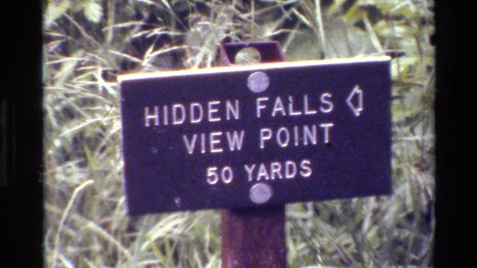 Dark Grand Teton trail sign reading Hidden Falls View Point 50 Yards stands in tall grass on 1980 8mm footage.