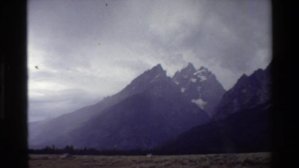 A cloudy Grand Teton landscape shows jagged peaks, snow patches, and a dark foreground in vintage 8mm texture.