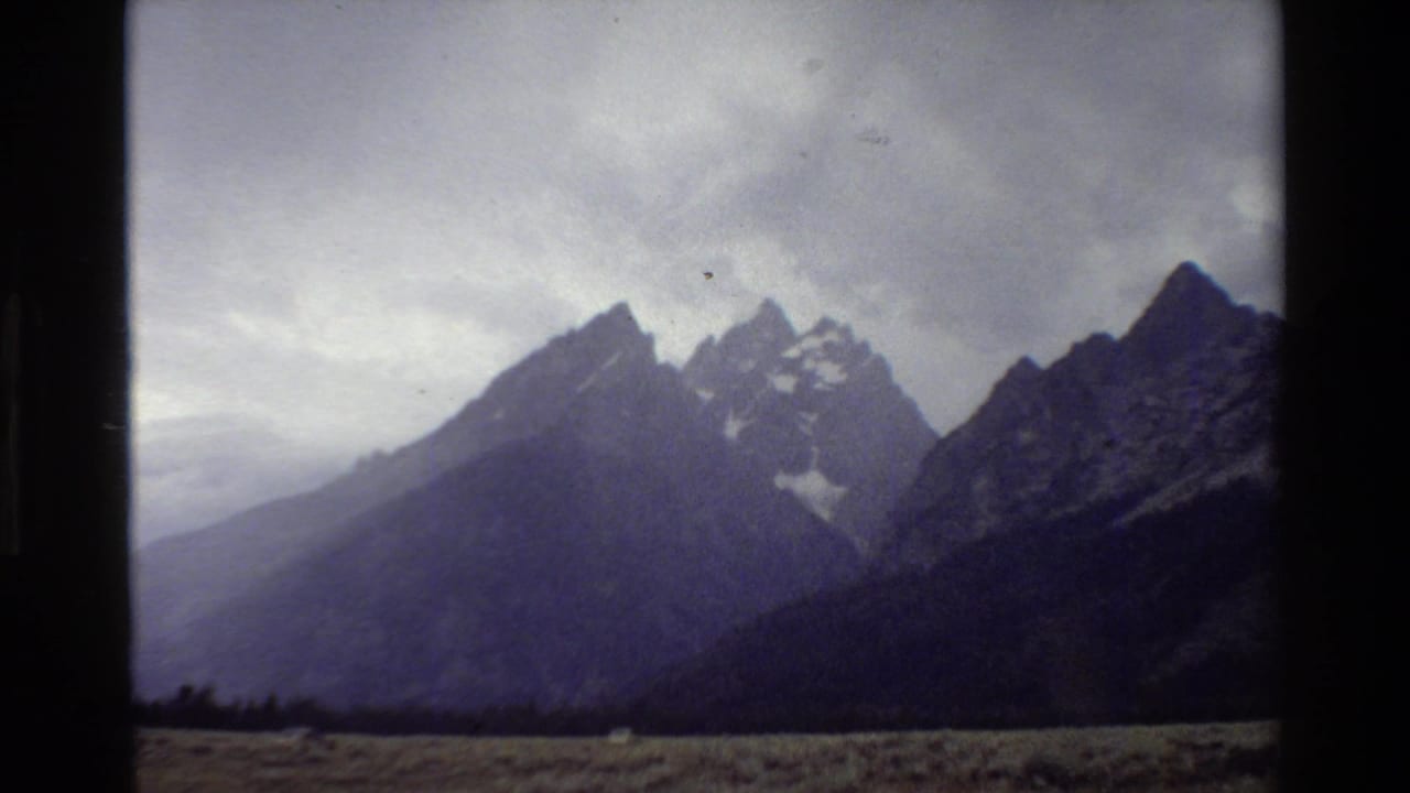 1980 8mm view of the Grand Teton range under overcast sky, seen across a grassy plain with dark film-strip edges.