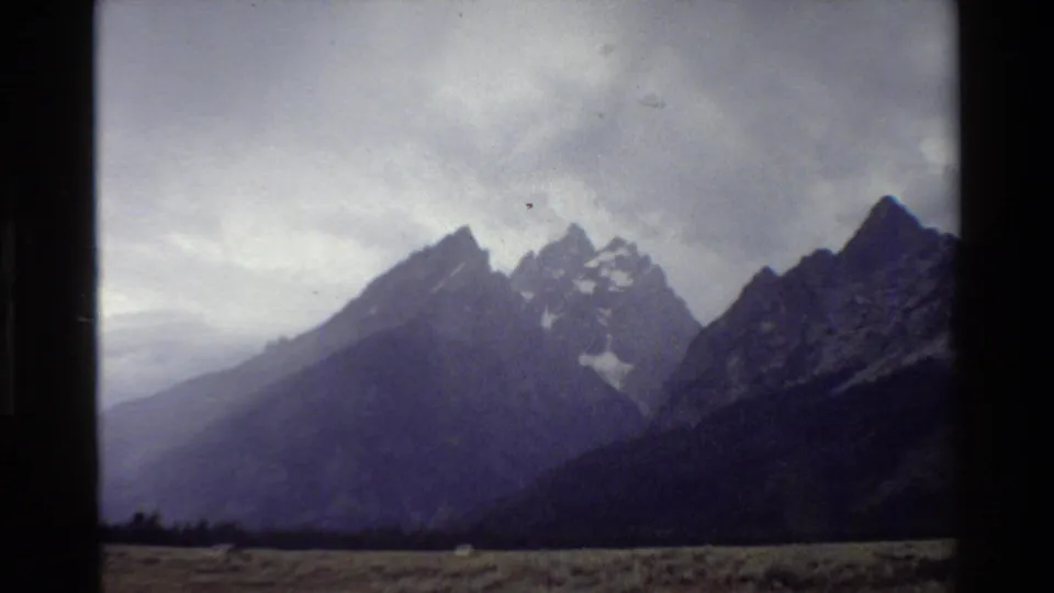 1980 8mm view of the Grand Teton range under overcast sky, seen across a grassy plain with dark film-strip edges.