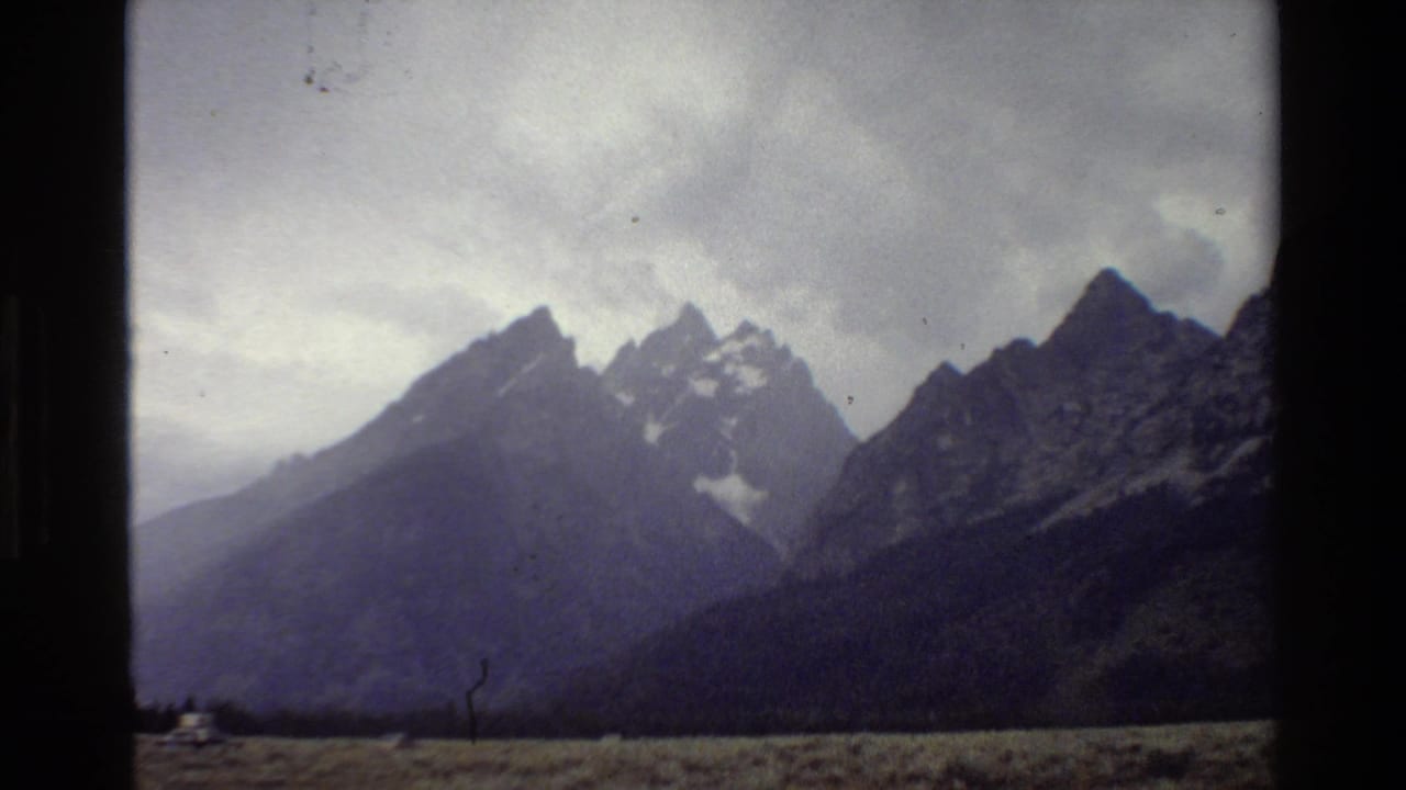 Mountain peaks and cloudy sky with grassy plain, dark frame edges, and a small pale shape near the horizon.
