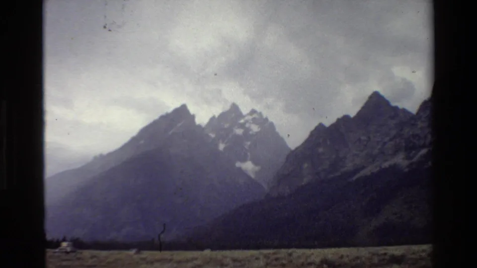 Grand Teton landscape in 1980 home-movie footage, with snow-marked peaks, cloudy sky, and a small distant roadside shape.