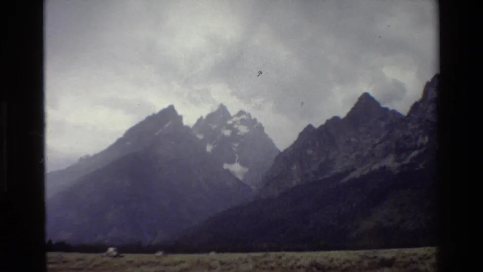 1980 8mm Grand Teton view with a small distant vehicle crossing the lower left edge of the grassy plain.