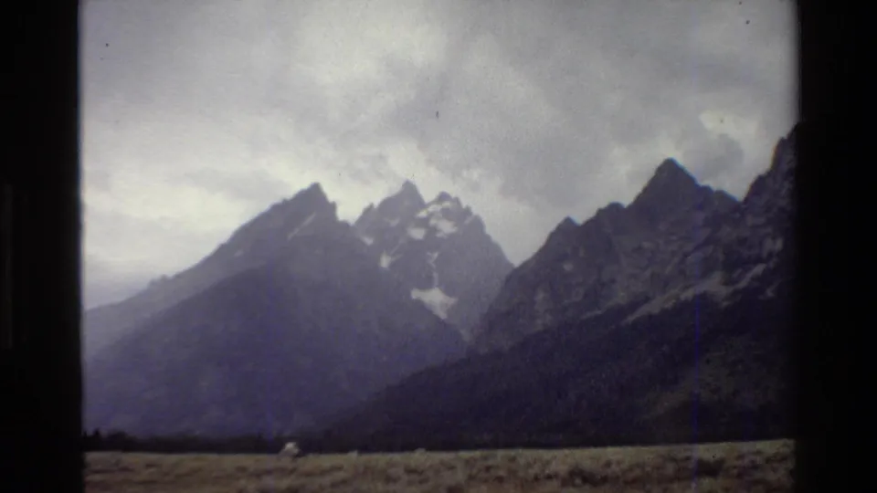 Soft 1980 8mm view of the Grand Teton range, with overcast sky, grassy plain, and a small pale roadside object near the bottom edge.