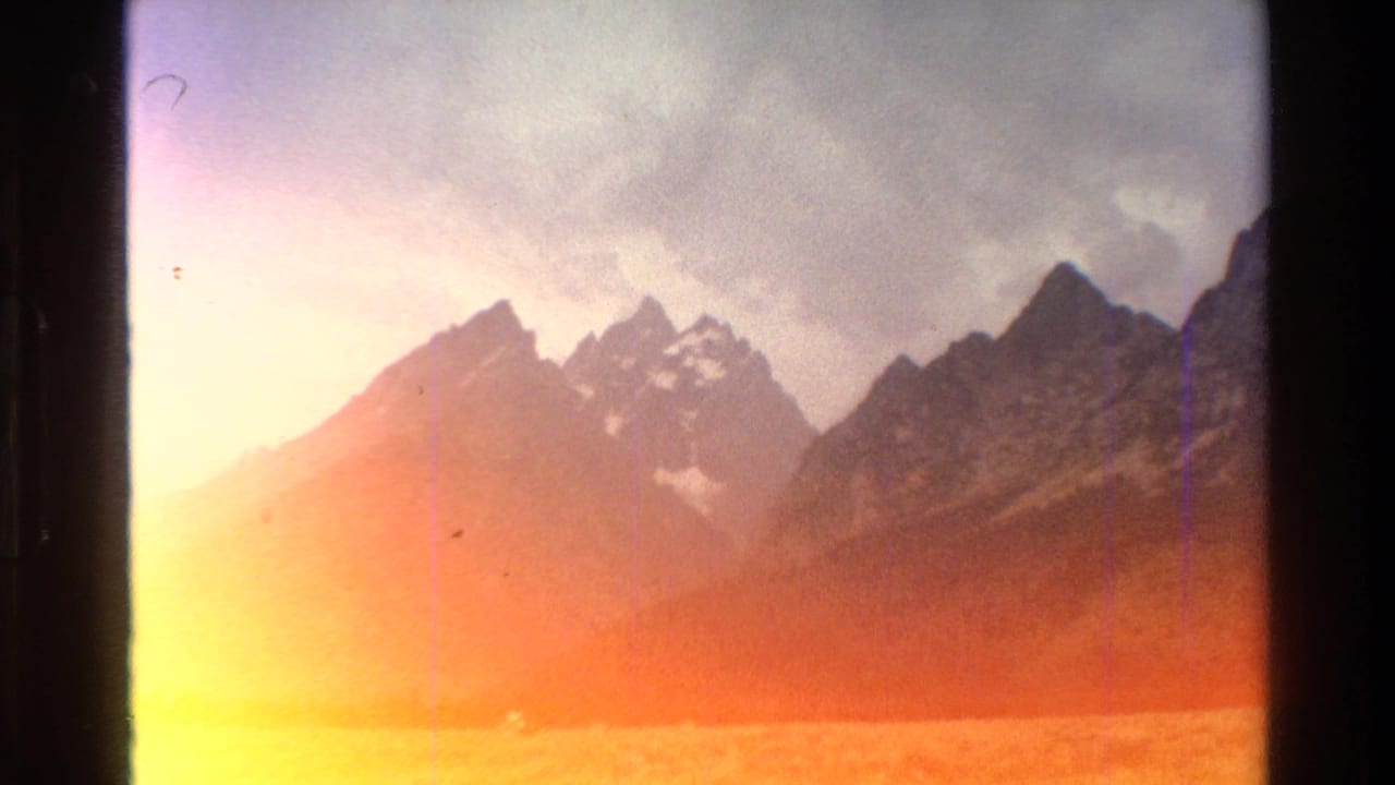 1980 8mm Grand Teton travel footage showing jagged mountain peaks, cloudy sky, and warm film flare near the bottom edge.