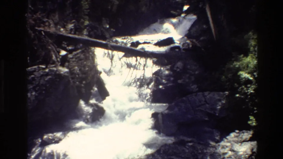 1980 8mm Grand Teton footage of a mountain stream cascading over dark rocks with branches and a fallen log above the water.