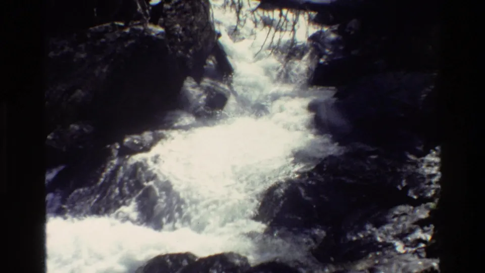 Grand Teton home-movie frame showing whitewater flowing between boulders in a shaded mountain stream channel.