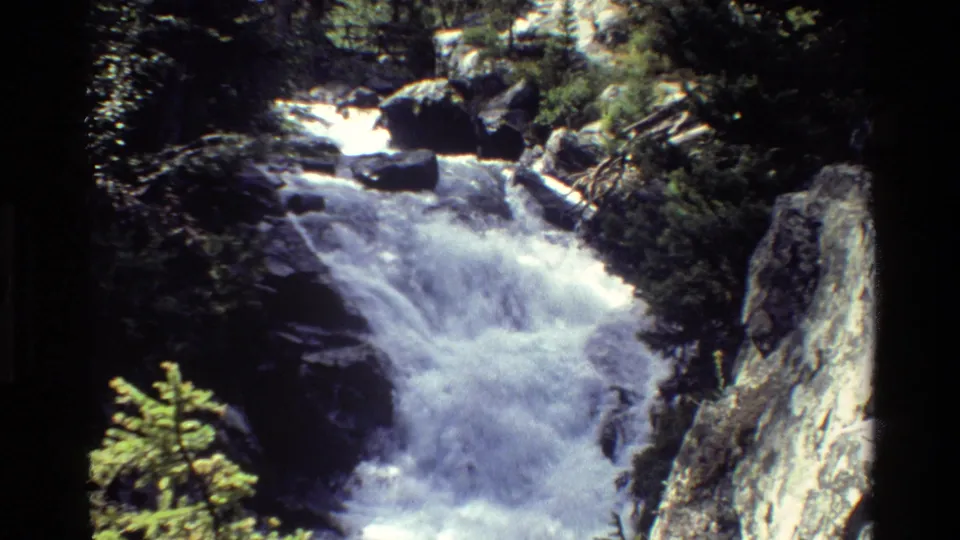 Grand Teton-area home-movie frame of bright rapids flowing between boulders and forest vegetation.