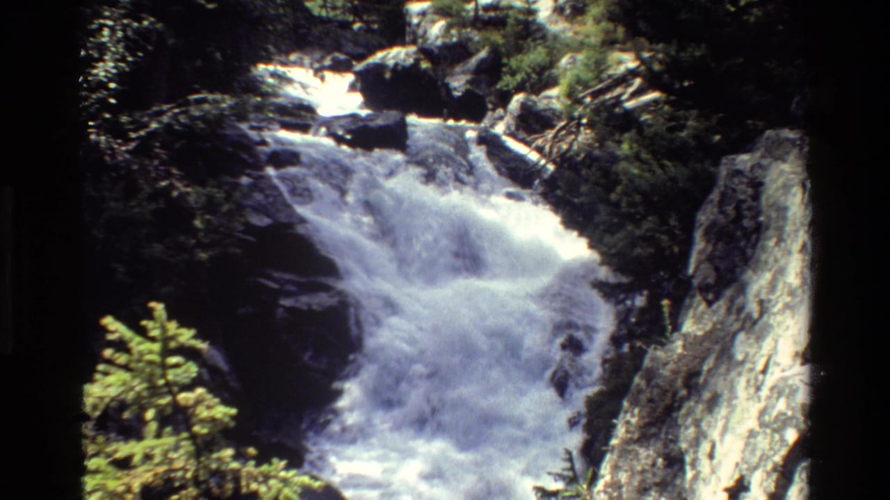 1980 8mm view of a Grand Teton waterfall cascading through rocks, framed by evergreens and a steep light-colored cliff.