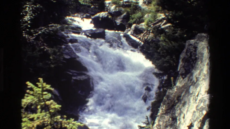 1980 8mm view of a Grand Teton waterfall cascading through rocks, framed by evergreens and a steep light-colored cliff.