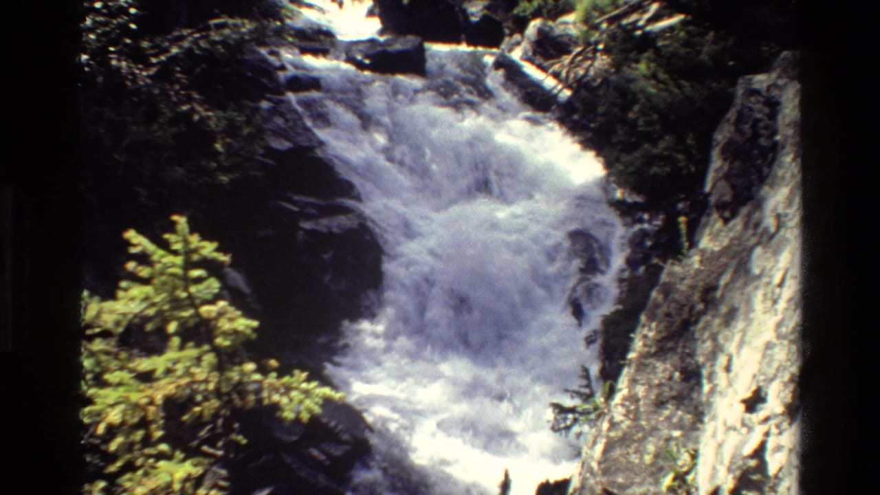 Foaming waterfall drops through a rocky chute with evergreens and a cliff at the side.