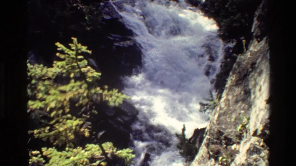 1980 8mm frame of a Grand Teton waterfall continuing downhill beside conifers and a rugged pale rock wall.