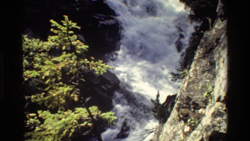 Final 1980 Grand Teton frame showing the waterfall spilling lower through rocks, with evergreens and a pale cliff beside it.