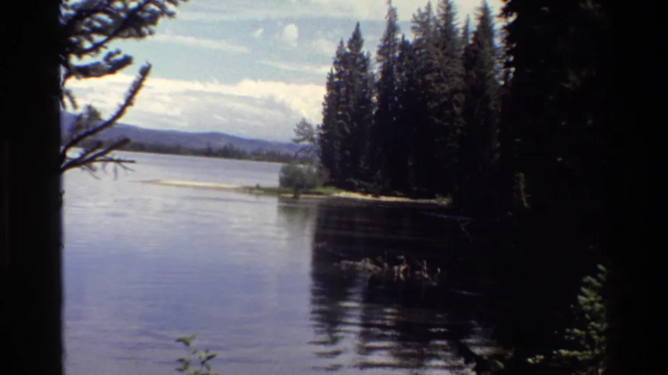 Tilted 8mm frame from Grand Teton showing a calm lake, evergreen shore, distant hills, and shaded foreground vegetation.