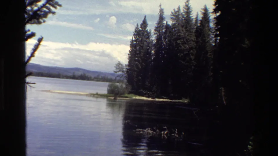 Grand Teton 1980 home-movie frame of a lake bordered by evergreens, with shaded trees, distant hills, and indistinct dark water shapes.
