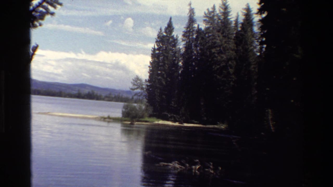 1980 Grand Teton home-movie frame showing calm lake water, dark conifers, distant mountains, and a partly cloudy sky.