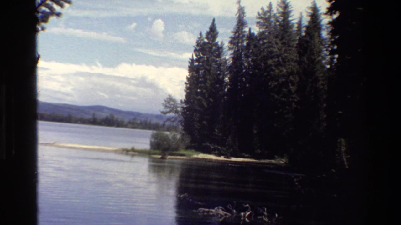Grand Teton lake scenery with conifer shoreline, distant hills, bright clouds, and visible 8mm frame borders.
