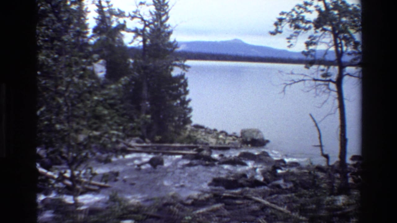 Rocky stream flows toward a calm lake with trees and distant mountains beyond.