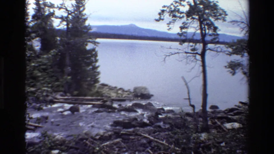 1980 8mm Grand Teton view of stream water entering a lake, framed by forest, shoreline rocks, and a distant ridge.