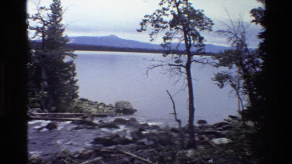 Grand Teton home-movie frame showing a creek mouth, calm lake, shoreline trees, and distant mountain forms under overcast light.
