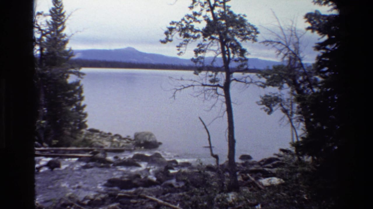 1980 Grand Teton home-movie frame showing a calm lake, rocky shore, evergreen trees, and distant mountains.
