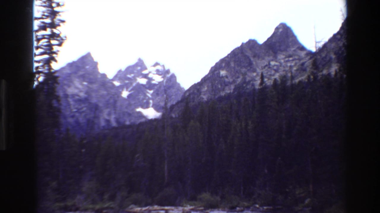 1980 8mm Grand Teton view with jagged peaks, small snow patches, evergreen forest, and dark film-frame edges.